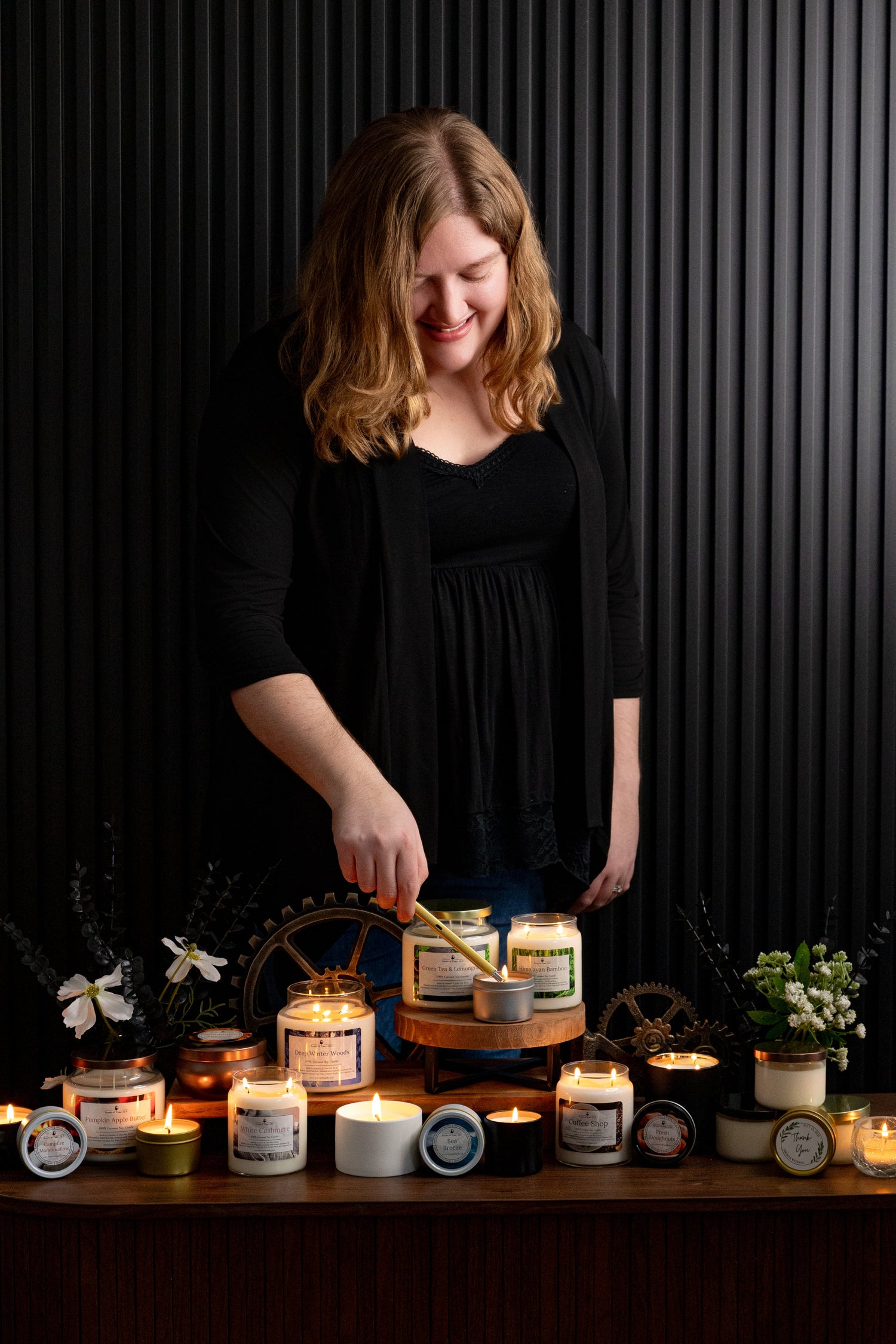 A picture of Katie, the founder of Scents of Time, with a black accent wall background. Katie is standing behind a variety of candles that she hand-poured in Lemont, IL. She is using an electric (eco) lighter to light a candle in the middle of the table. Most of the candles are lit in the photo.