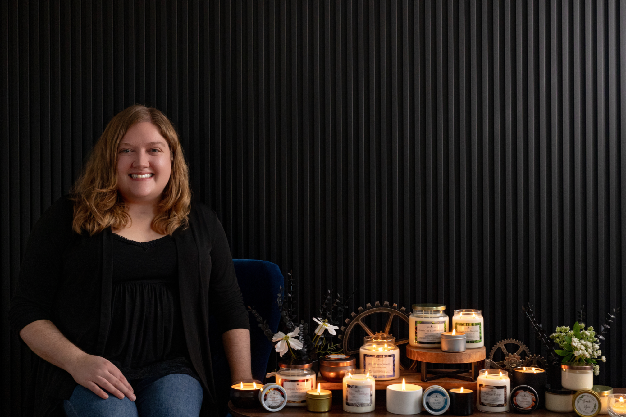 A picture of Katie, the founder of Scents of Time, with a black accent wall background. Katie is sitting next to a variety of candles that she hand-poured in Lemont, IL. Most of the candles are lit in the photo.
